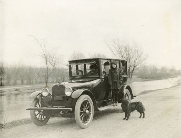 Dick Lloyd Jones stands on the running board of the family car parked along the Yahara River Parkway in Tenney Park.  Blig, the family's Belgian Shepherd dog, stands alongside. Two women and two children are sitting in the car.