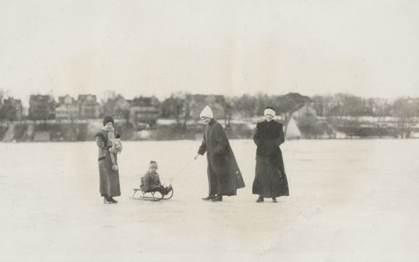 A woman pulls Richard "Dick" Lloyd Jones on a sled across the ice on Lake Monona. Two other women stand on either side, and houses on the shoreline are in the background.