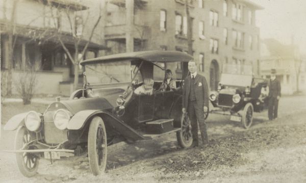 Unidentified man stands beside a car, with Richard "Dick" Lloyd Jones and Jenkin Lloyd Jones inside.  An unidentified man poses beside a second car.  In the background is the Monona Apartments building at the corner of South Brearly and Spaight Streets.