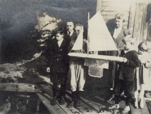 A group standing on a Lake Monona pier with a large toy sail boat.  Richard "Dick" Lloyd Jones is at far left, his mother, Georgia Lloyd Jones stands to the right of the boat, behind her two other children, Jenkin and Florence (Bis).  Two other people are unidentified.
