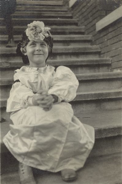 Florence (Bis) Lloyd Jones sitting on steps in a fancy dress, high button shoes, and a hat with a large flower.