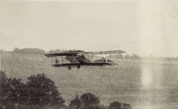 A Curtiss JN-4 "Jenny" airplane / biplane in flight over the Weber Farm south of Lake Monona.  Titled in the album "The First Aeroplane in Madison". "The feature of the flight was the delivery of a letter to Postmaster Devine from Pres. William F. Brooks of the Minneapolis Aero Club.  This is the first aerial letter ever received in Madison and the first aerial postage stamp ever received here.  The flyers are returning from a 3,000 mile run and have been making exhibition flights at state fairs."  The Capital Times 9-4-1918.  One of five airplane images taken over a two day period.