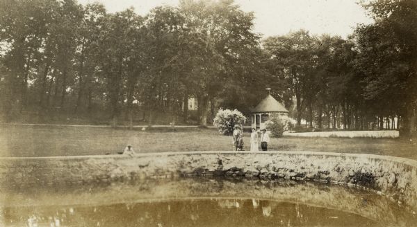 A woman and children stand beside a pond at the Wisconsin State Fish hatchery (aka Nevin Fish Hatchery, 3911 Fish Hatchery Road) south of Madison. A gazebo or pavilion is in the background.
