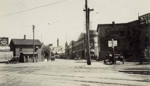 A view east from the corner of South Blair and Williamson Streets.  On the left is Joe's News Store (Joseph J. Kuhn) 520 E. Wilson Street and a sign for the Auto Shop, 606 Williamson Street. On the right (south) side of Williamson Street are the Fauerbach Brewery, 651 Williamson Street, Machinery Row, 601-631 Williamson Street, Avery Co., 521-523 Williamson Street and Nuss Implement Co., 513 - 519 Williamson Street. A manure spreader is parked near a railroad crossing sign in front of the Nuss Implement Company.