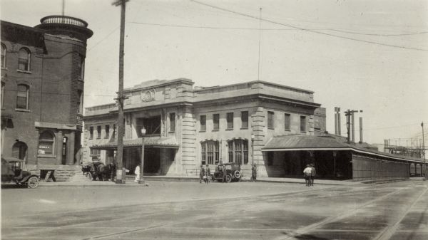 A view of Chicago & North Western Railroad Station, 219 South Blair at East Wilson Street, which was built 1910.  The Elver House hotel, 520-524 East Wilson Street, is on the left.  Automobiles and a horse-drawn carriage are parked along the street.