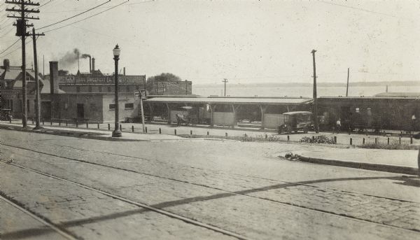 The East Madison Chicago, Milwaukee, and St. Paul railroad station, 501 East Wilson Street, from South Hancock Street, with Lake Monona in the background.  A train is stopped at the station, and a bus and horse-drawn carriage are parked near the platform.  The Nuss Implement Co. is behind the station.