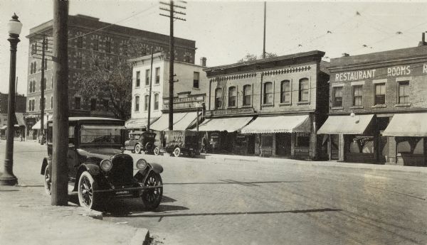 A view looking west up the 500 block of East Wilson Street.  Left to right are the Cardinal Hotel, 416-18 East Wilson Street; Lake City House, 502 East Wilson Street; Louis Russos candy store, 504 East Wilson Street; Herman Klueter grocery & real estate, 506 East Wilson Street; East Madison Market H.A. Vogt, 508 East Wilson Street; Lake View Hotel and Quick Lunch, 510 East Wilson Street. There are also two automobiles, one with a trailer, in the foreground.