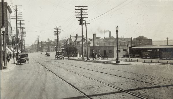 A view looking east along East Wilson Street with the Chicago, Milwaukee, and St. Paul East Madison railroad station, 501 East Wilson Street, on the right. Several pedestrians and automobiles are also visible. Lake Monona is in the background behind the railroad station.