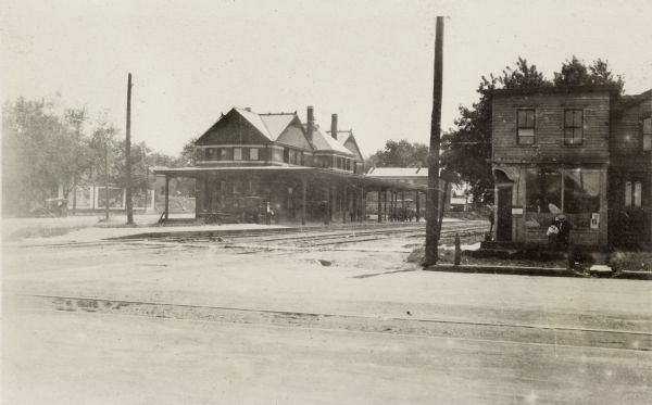 Illinois Central Railroad station.  Automobiles and a horse-drawn wagon are parked beneath the canopy.  In the right foreground is a commercial building with a man and boy looking in the window of the storefront.