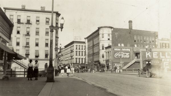 Street scene with pedestrians near Milwaukee Street bridge.  The Hotel Myers is in the background.  The Lappin-Hayes Block is seen to the right of the Myers Hotel.  There are several billboards on the sides of buildings, including one for Coca-Cola. Richard Lloyd Jones was born in Janesville in 1873 and lived there until his father, Rev. Jenkin Lloyd Jones moved the family to Chicago in 1881. This picture was taken when Richard moved his family from Madison to Tulsa, Oklahoma, in 1919.