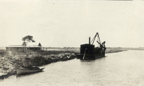 The dredge used for clearing the channel of the Yahara River between Lakes Monona and Waubesa.  Two cottages are on the shore and a rowboat is tied along the bank.