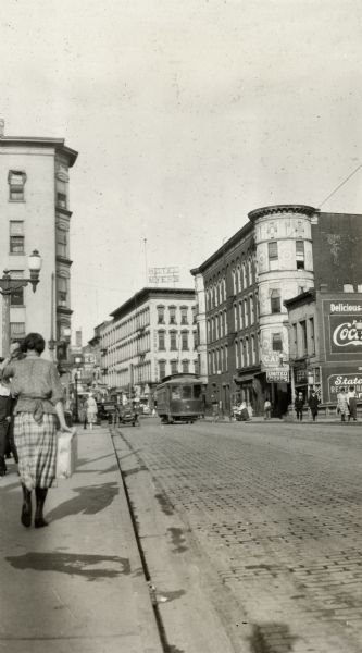 Pedestrians in shopping district. Cars are parked and a streetcar is coming down the street.