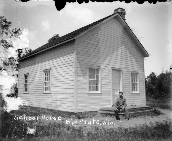 Exterior of the schoolhouse with man sitting on front steps.