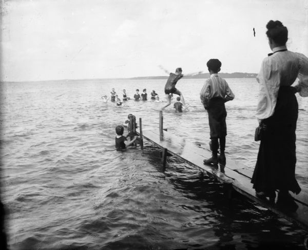 Children swimming and jumping off pier into lake. An adult woman appears to be supervising the children.