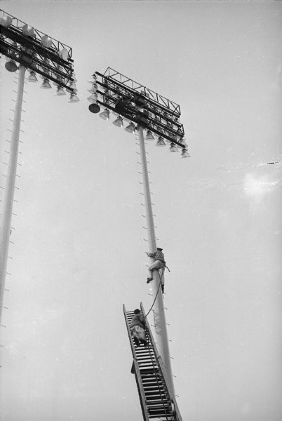 Milwaukee County Stadium on the day of the first game of the 1958 World Series, between the Milwaukee Braves and the New York Yankees. A man is climbing the stadium lights to reach another man already atop the lighting platform.
