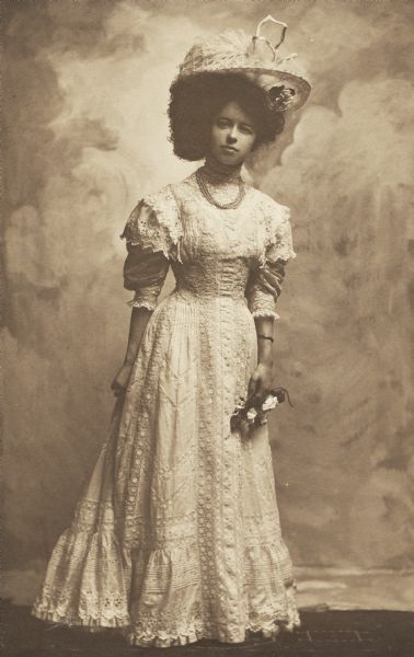 Full-length studio portrait of Bettina Jackson, of Madison, wearing the dress that she wore in the wedding of her brother Reginald to Elsie Stevens in 1908. The dress is described as lace and embroidery over pale pink, the hat of pink tulle with an ostrich plume.