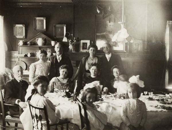 Informal portrait of the W.A.P. Morris family, with the patriarch seated at the head of the table at the left, and his children and grandchildren.  Portraits of his forebears, including Revolutionary War General Jacob Morris, his father, hang on the wall.  The two men standing in the back row have been tentatively identified as Morris' sons: A. Breese Morris and Charles M. Morris (right).  Charles stands next to his wife, Mary Fairchild Morris, the daughter of Lucius Fairchild.  A print of the Fairchild portrait by John Singer Sargent can be seen on the mantle, along with a photograph of Reuben Gold Thwaites, head of the Wisconsin Historical Society and a close family friend.