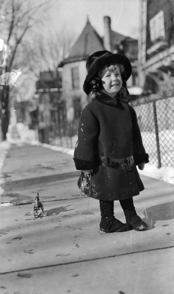 Reginald Jackson, Jr. is posed on the sidewalk in front of the family home, 415 North Carroll Street, dressed in his winter coat. The portrait is notable for young Reginald's curls and feminine-looking attire.  
