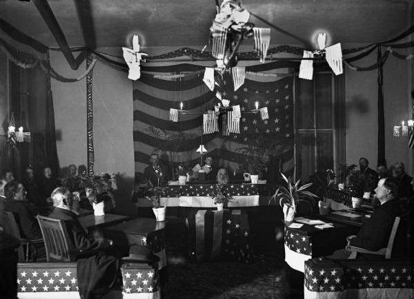 Men gather at lodge hall, probably in Watertown, Wisconsin. American flags are hanging from the light fixtures, draped over tables and on the wall. There is a man sitting with his head just above a hole in a table at the front of the room. The other men are sitting in chairs in front of small tables around him.