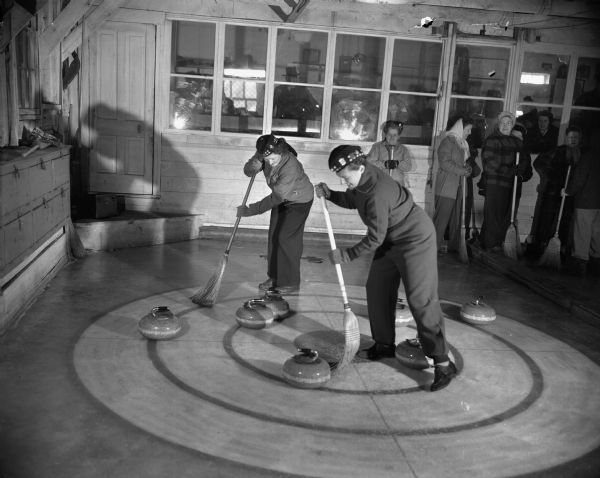Two women compete in a curling match. Several spectators are visible in the background.