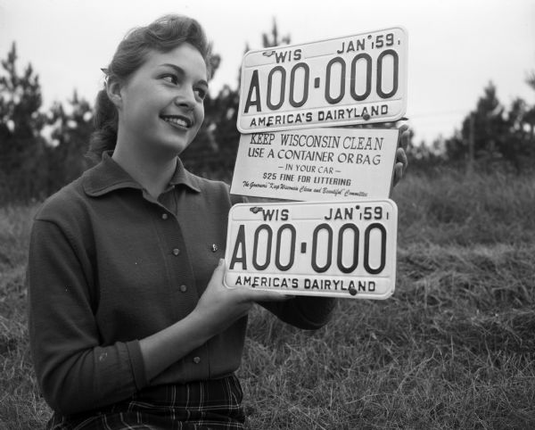 A young woman standing outdoors holds two 1959, Wisconsin license plates and a "Keep Wisconsin Clean" sign. The signs were made to promote the Governor's Keep Wisconsin Clean and Beautiful Committee.
