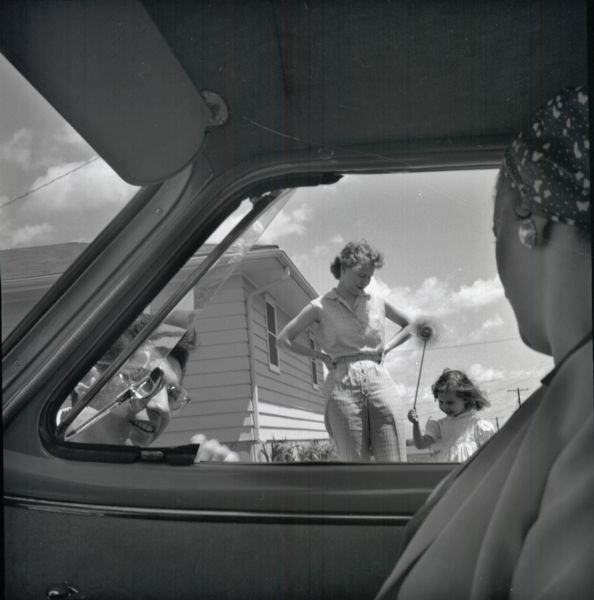 View from the interior of a car looking out of the passenger window. A woman is sitting on the passenger side of the car. A boy standing outside is looking into the car, and in behind him is a woman standing with her hands on her hips beside a small girl who is holding a pinwheel. There is a house in the background.