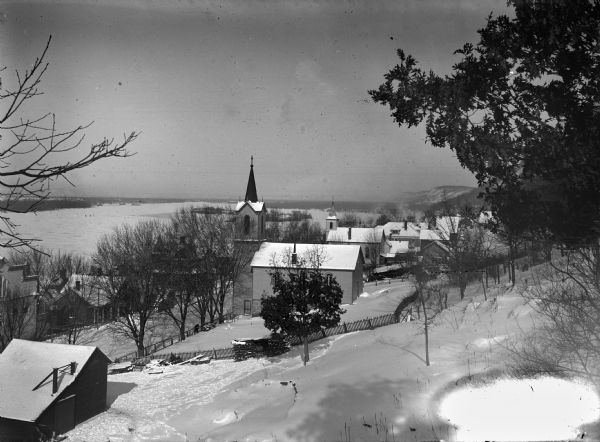 A view from a hill overlooking the town of Alma, on the banks of the Mississippi River, which is frozen. Several churches and residential houses are visible.