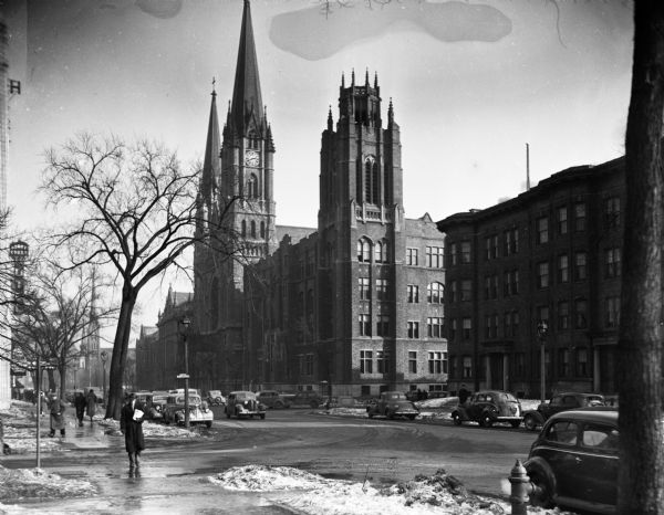 Gesu Church, with automobiles and pedestrians on West Wisconsin Avenue.  There is a sign on the left for the Abbot Crest Hotel located at 1226 West Wisconsin Avenue. The photograph was taken for the Writer's Program to be used as an illustration for <i>Wisconsin: a Guide to the Badger State</i>.