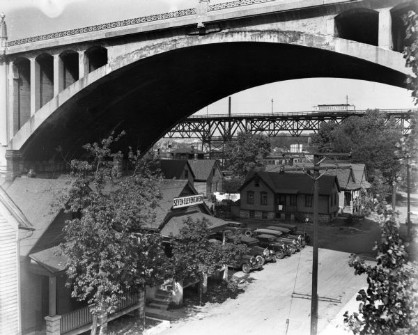 View of the Grand Avenue viaduct and surrounding establishments. A line of automobiles are parked outside a tavern under the viaduct.