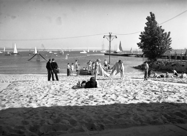 A beach on Lake Michigan near the Milwaukee Yacht Club. Men, women, and children relax on the beach. In the background many boats are on the water.