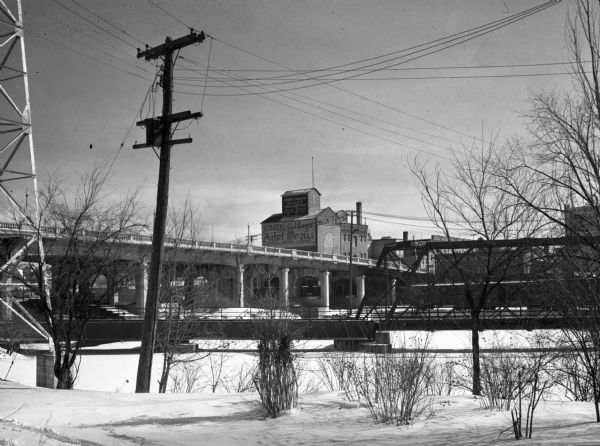 An exterior view of the Cereal Mills Company, located at 112-118 West Scott Street. A railroad and a highway bridge are in the foreground.