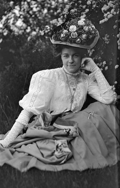An outdoor portrait of Mary E. Smith, taken by her husband Dr. Joseph F. Smith. Mrs. Smith is sitting in the grass, and wears an elaborate hat decorated with roses, and long gloves.
