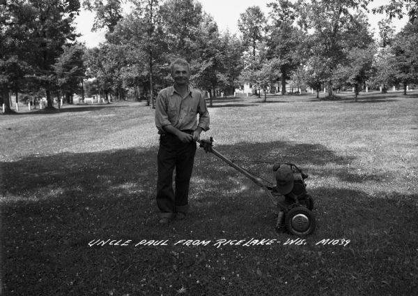 A man stands outdoors with a lawn mower. His work gloves and hat are resting on the mower. Caption on photograph reads, "Uncle Paul from Rice Lake-Wis."
