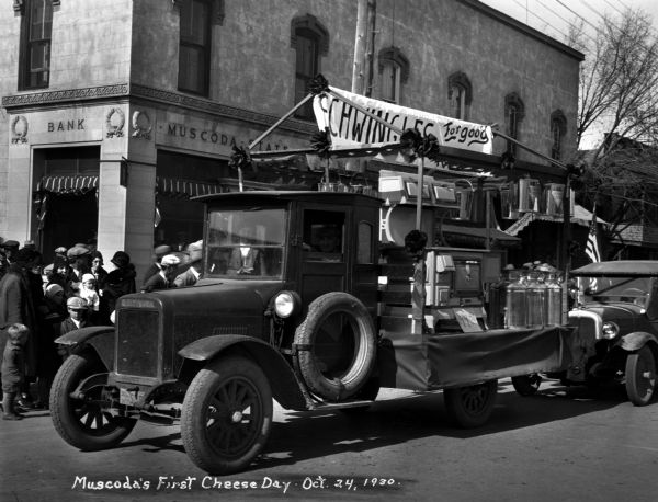 Celebrating the first "Cheese Day" with a parade. A group of spectators gather on the sidewalk near a truck displaying a banner and stove in the back.