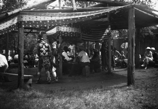 A small crowd gathers for a Winnebago (Ho-Chunk) ceremony under an open air structure, probably in the Wisconsin Dells. Two men in ceremonial dress stand in the foreground while others sit on benches in a circle watching.