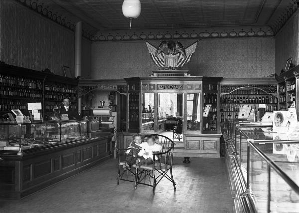 An interior view of Brennecke and Bergmann Pharmacy on the corner 4th and Main Street. Two children are sitting on a chair in the middle of the floor, while a man is standing behind the counter. A large built-in cabinet with mirrors is in the background, and a spittoon is on the floor. For many years Bergman's sideline was postcard photography.