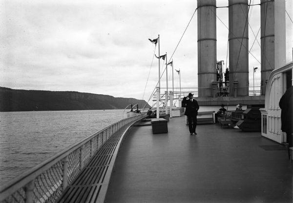 A man is standing on the deck of a large ship as it travels the Hudson River. Other passengers are sitting on benches in the background.