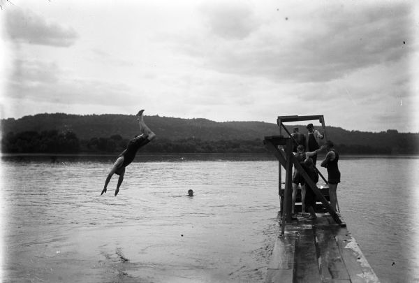 A man diving off a pier into the Mississippi River. Another man is swimming in the water, while other men and boys are standing on the floating pier.