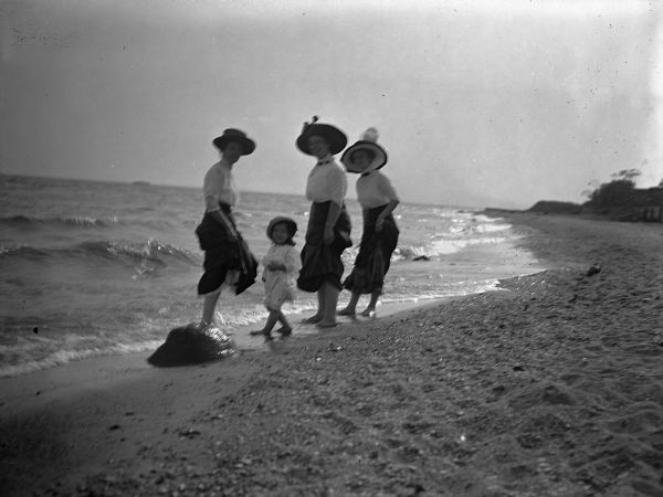 Three women and a child are wading at the Lake Park shoreline. The women are identified as Hattie Taylor and friends, and the child as Tannisse Taylor.