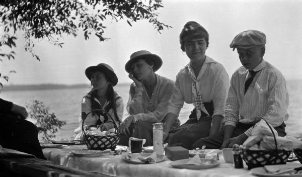 Four individuals, sitting in the shade near a table during a picnic on Madeline Island. Includes Elizabeth Nesbet (Mrs. S.C. Marty, Sr.), Gertrude Capser (Mrs. Gale Holland) and William Baker.