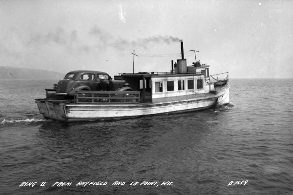 A car ferry transporting a car on Lake Superior between LaPointe, Madeline Island, and Bayfield. Two men are sitting on a bench near the car, and a shoreline can be seen in the background.