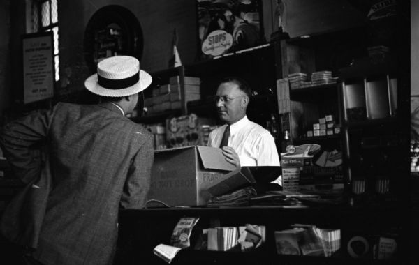 Two men talking in an auto parts store. One man is behind the counter wearing eyeglasses and holding a box, the other man is wearing a hat and leaning on the counter.