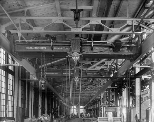 Interior view of two early 6-ton Pawling & Harnischfeger I-Beam cranes, numbered 123 and 124. The bridges of both are an early "O" type used for short spans. The stencil on both cranes read "Pawling and Harnischfeger, Milwaukee, Wis. Capacity 12,000 Lbs." with the nearest numbered "123" and the farther numbered "124." There are two pieces of machinery along the right side of the picture that are stamped "The Pond Machine Tool Co., Plainfield, NJ, U.S.A."