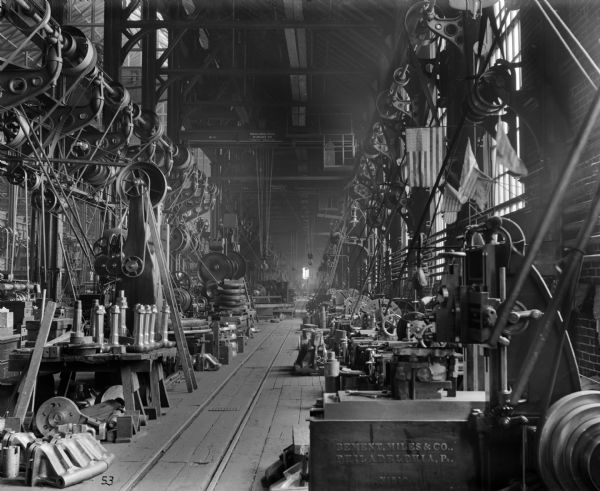 Interior view of a machine shop outfitted with two early Pawling & Harnischfeger 15-ton, overhead trolley, cab-operated, basic block I-Beam cranes with type "O" bridges and type "A" trolleys. Both cranes are stamped "Pawling and Harnischfeger, Milwaukee, Wis. Capacity 20,000 Lbs." with the nearest crane numbered "110" and the farther crane numbered "111." In the foreground to the right is a piece of machinery stamped "Bement, Miles & Co., Philadelphia, PA No 510." There are American Flags visible along the right wall of the machine shop.