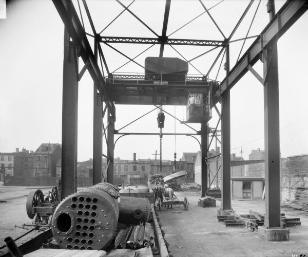 Pawling & Harnischfeger 40-ton type "A" outdoor gantry crane located at the Pennsylvania Railroad Co. There are men standing near a horse-drawn cart as well as a man standing at the door to the cab of the crane. The crane is stamped (though partially obscured) "Pawling & Harnischfeger Builders, Milwaukee, Wis., No. 563." The building in the background is identified as "Mason & Builders. Designers. Of All Kinds Of Firework & Furnace. Cremating Furnaces & Factory Work Of Every [obscured]." The building in the background on the right reads "S.B. Vrooman Co. Ltd. [unreadable] Mahogany & Hardwood."