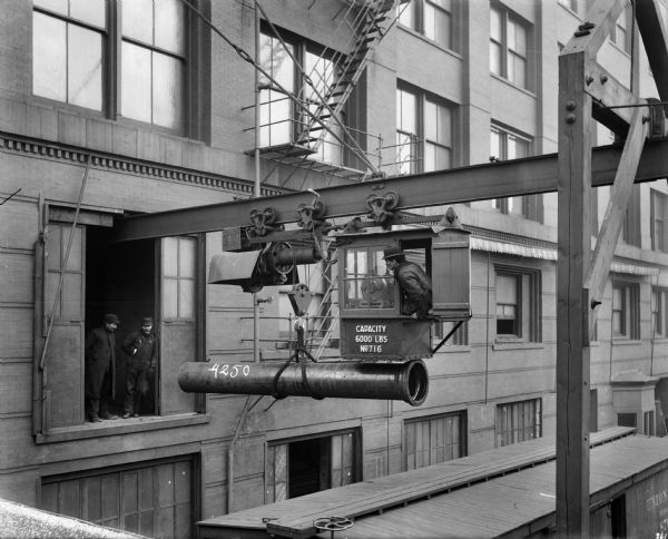 Exterior view of a Pawling & Harnischfeger 3-ton traveling electric monorail hoist located at James B. Clow & Sons. The cab is stamped "Capacity 6,000 Lbs., No. 716." There is an operator in the cab, and two men are standing in an open doorway, which is above a railroad car.