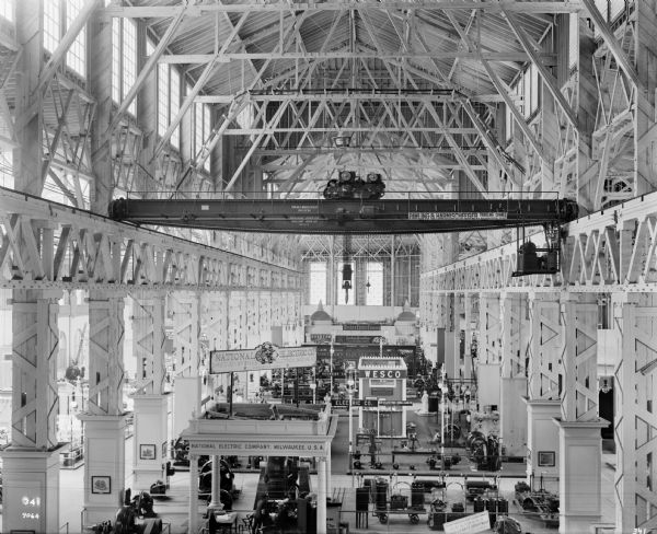 Pawling & Harnischfeger 30-ton type "AN" bridge crane with a 5-ton type "A" trolley at the 1904 St. Louis World's Fair in either the Palace of Electricity or Machinery. The crane is stamped with the text "Pawling & Harnischfeger Builders, Milwaukee, Wis., Main Hoist 60,000 Lbs., Auxiliary 1,000 Lbs., No. 1075." and the banner draped on the crane reads "Pawling & Harnischfeger, Traveling Cranes, Milwaukee, Wis." There are booths from various companies on the floor of the room.