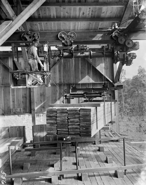 Pawling & Harnischfeger monorail hoist with a lumber handling unit, laden with lumber, with operator repositioning load as the hoist leaves the lumber shed through a large opening.