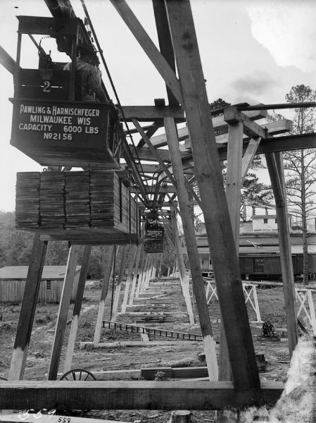 Rear view of two Pawling & Harnischfeger 3-ton monorail hoists with lumber handling units laden with lumber and men in the cabs. Both hoists  are stamped with text that reads "Pawling & Harnischfeger Builders, Milwaukee, Wis., Capacity 6,000 Lbs." with the nearest hoist identifed as "No. 2156" and the farther one "No. 2155."