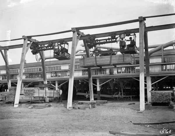 Two Pawling & Harnischfeger 3-ton monorail hoists with lumber handling units laden with lumber and men in the cabs. Both hoists are stamped with text that reads "Pawling & Harnischfeger Builders, Milwaukee, Wis., Capacity 6,000 Lbs." with the hoist on the left identifed as "No. 2155" while the hoist on the right has its number obscured.